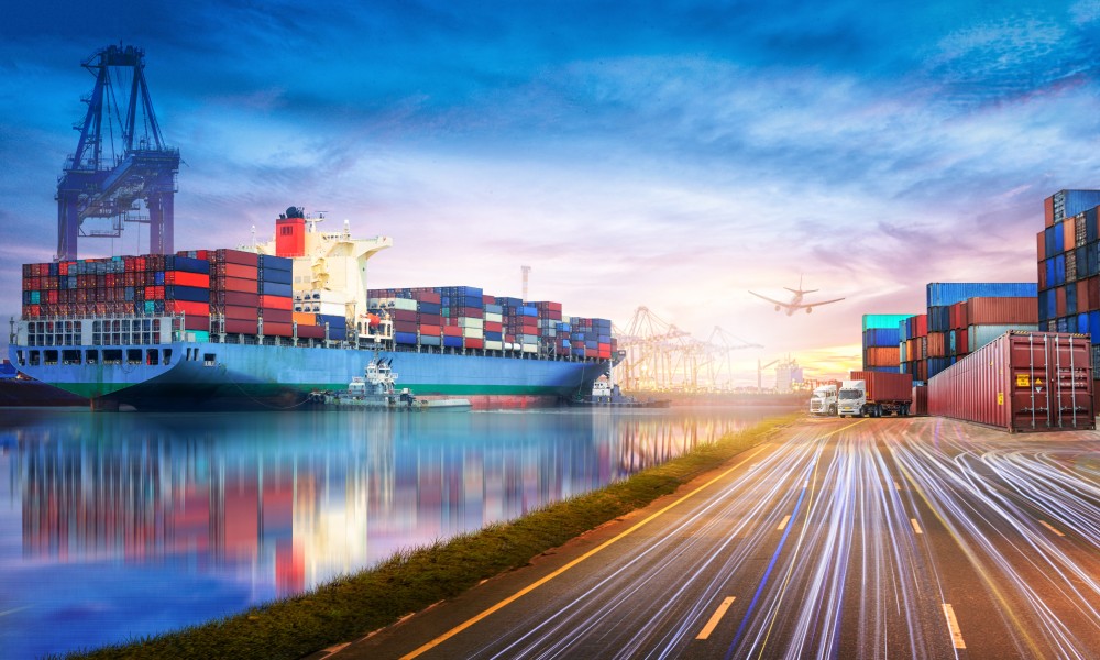 A fully loaded cargo ship in the ocean next to a busy road and a freight plane flying overhead.