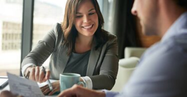 A woman in a blazer smiling as she reaches over a table to point to something on a paper her male client is holding.