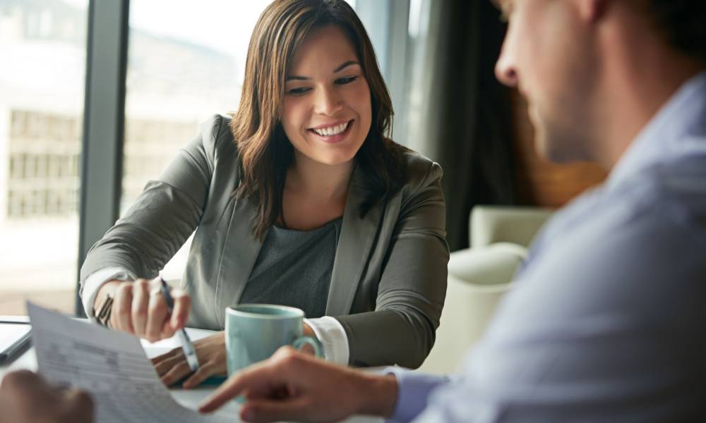 A woman in a blazer smiling as she reaches over a table to point to something on a paper her male client is holding.