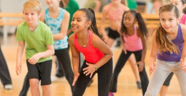 A group of diverse children engaged in a dance class, joyfully participating and interacting with each other. The focus is on a young girl in a red top, expressing her happiness and confidence while dancing.