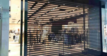 A secure roll-down iron gate guards the entrance of a retail storefront. The lights are still on inside the store.