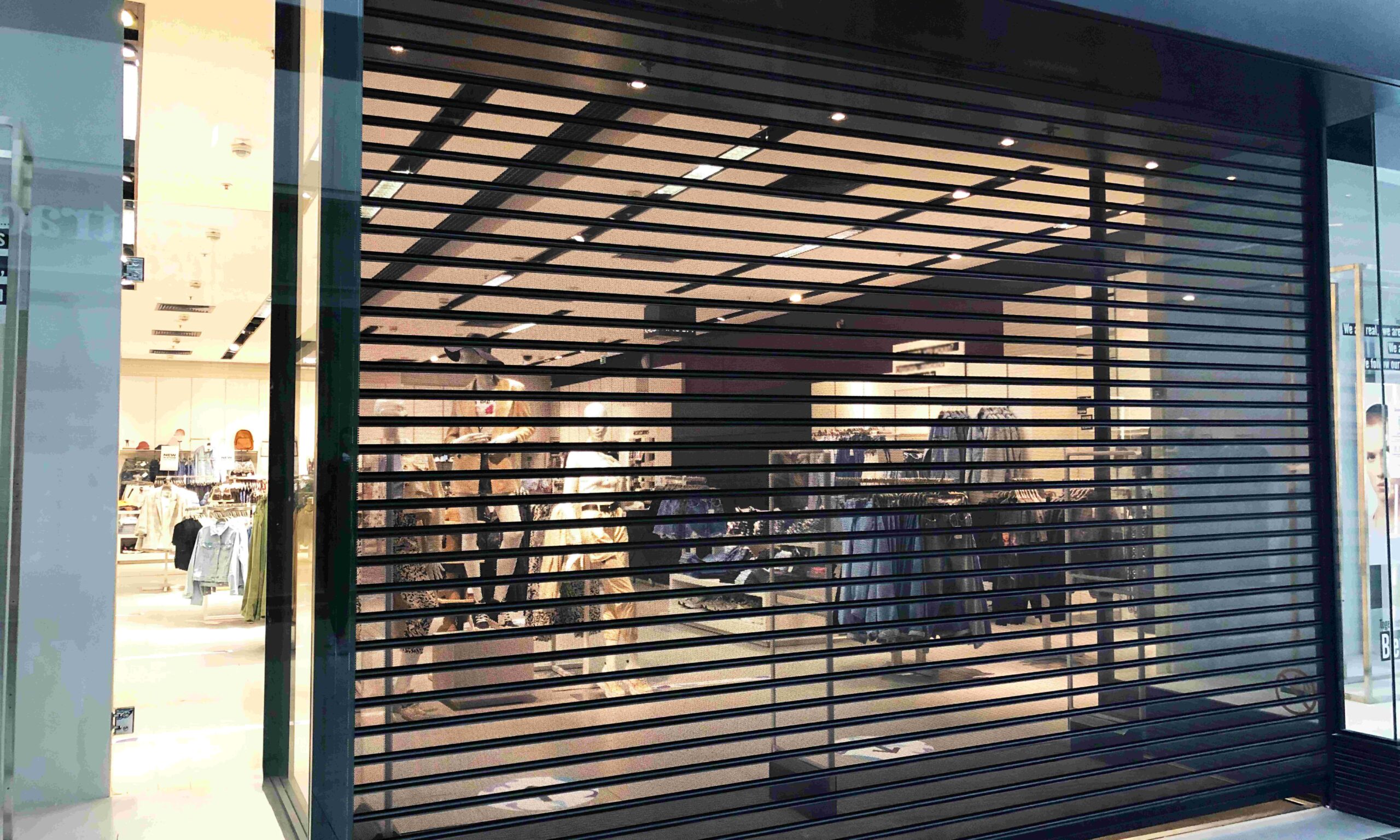 A secure roll-down iron gate guards the entrance of a retail storefront. The lights are still on inside the store.