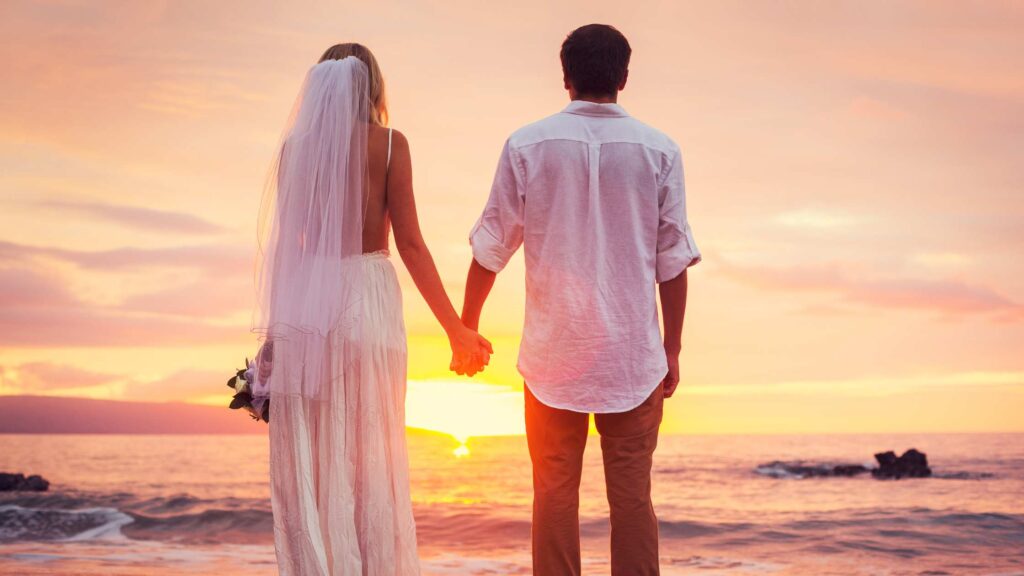 A couple stands hand-in-hand, facing a breathtaking sunset on the beach on their wedding day. The bride in a flowing veil and the groom in a casual white shirt capture a moment of romance and anticipation against a vibrant sky.