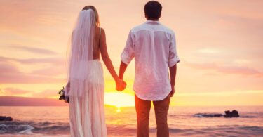 A couple stands hand-in-hand, facing a breathtaking sunset on the beach on their wedding day. The bride in a flowing veil and the groom in a casual white shirt capture a moment of romance and anticipation against a vibrant sky.