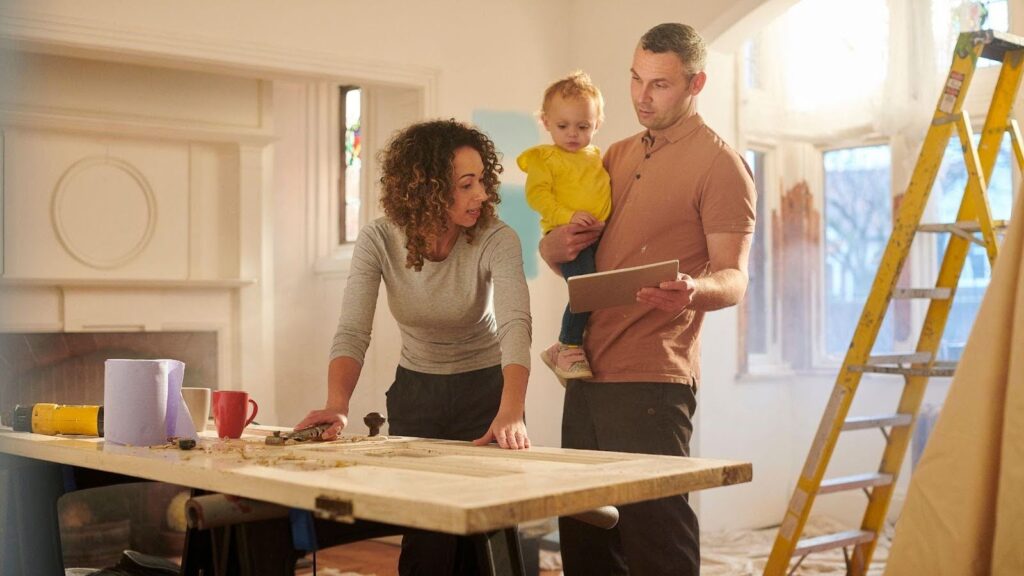 A family engaged in a DIY home improvement project in a brightly lit room under renovation. The mother, with curly hair, leans over a worktable examining tools, while the father, holding a toddler and a digital tablet, discusses plans. 