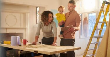 A family engaged in a DIY home improvement project in a brightly lit room under renovation. The mother, with curly hair, leans over a worktable examining tools, while the father, holding a toddler and a digital tablet, discusses plans.