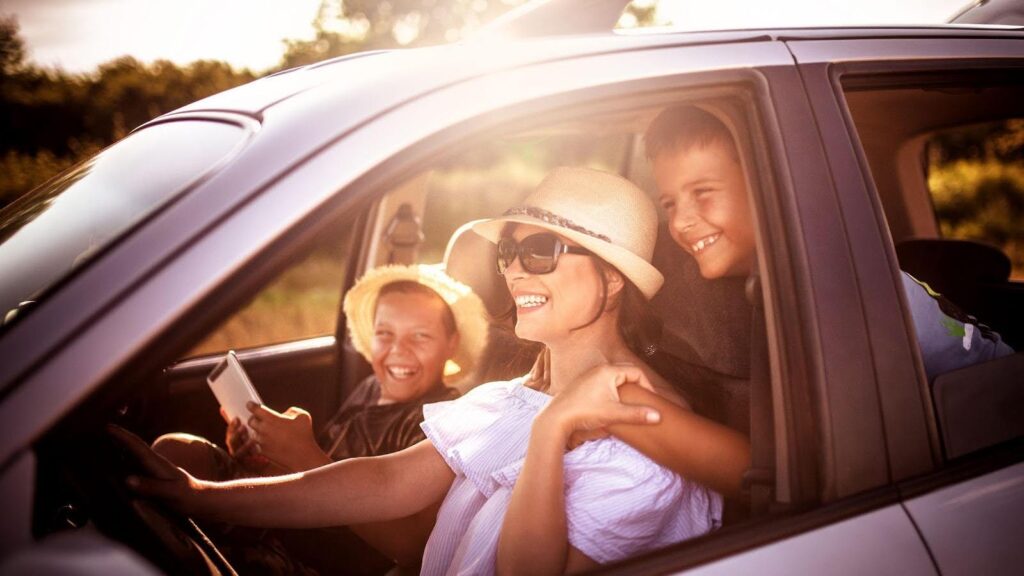 A joyful family in a car during a sunny road trip, featuring a mother in sunglasses and a straw hat driving, with her two laughing children in the passenger seats, one holding a tablet. 