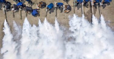 Eighteen workers aligned holding chemical foggers in an outdoor environment spraying at the same time creating a big cloud.