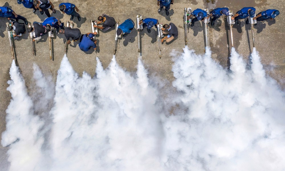 Eighteen workers aligned holding chemical foggers in an outdoor environment spraying at the same time creating a big cloud.