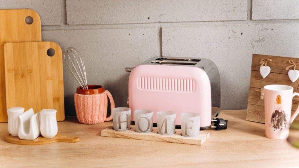 Cosy kitchen counter displaying vintage kitchen appliances including a pastel pink toaster, surrounded by wooden cutting boards, ceramic creamers, and a rustic 'HOME' decoration, illustrating how classic elements can enhance the aesthetic value of a home. 