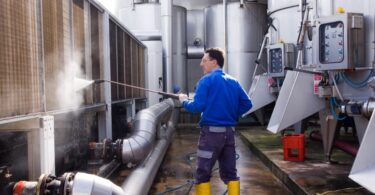 A man with glasses in a blue shirt and pants uses a high-pressure washer to clean off large machinery.