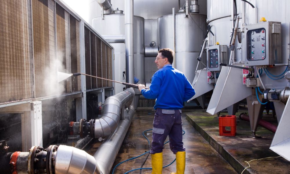 A man with glasses in a blue shirt and pants uses a high-pressure washer to clean off large machinery.