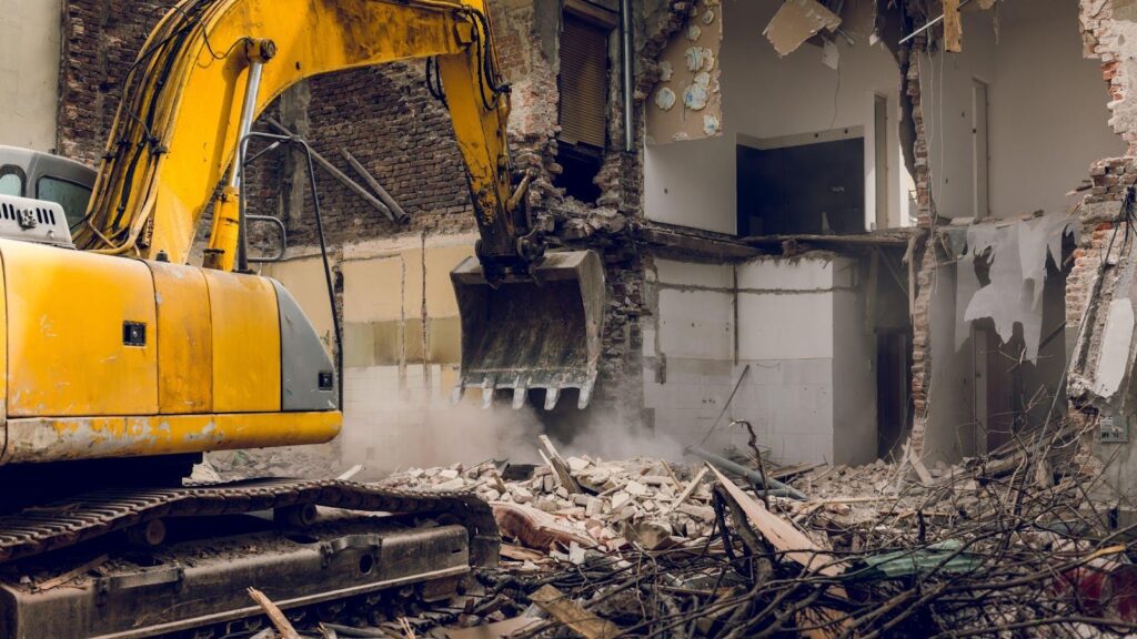 An excavator at work in a partially demolished building after a fire incident, tearing down walls amidst a cloud of dust. The scene shows debris and exposed structural elements, illustrating the complex and hazardous nature of safe demolition practices necessary in the aftermath of a fire.