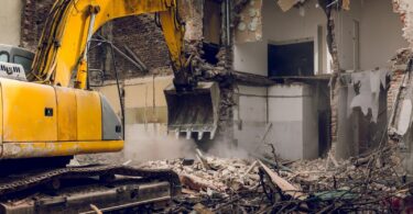 An excavator at work in a partially demolished building after a fire incident, tearing down walls amidst a cloud of dust. The scene shows debris and exposed structural elements, illustrating the complex and hazardous nature of safe demolition practices necessary in the aftermath of a fire.