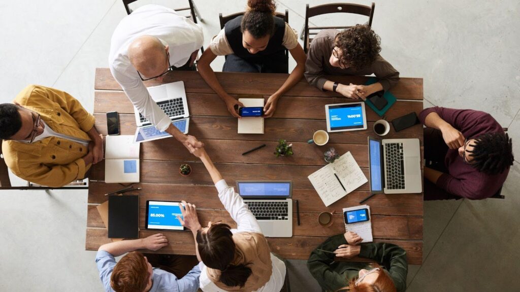 A group of office workers sitting around a large wooden table, working on laptops and tablets, while two colleagues shake hands, symbolising collaboration and productivity in a professional setting.