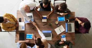 A group of office workers sitting around a large wooden table, working on laptops and tablets, while two colleagues shake hands, symbolising collaboration and productivity in a professional setting.