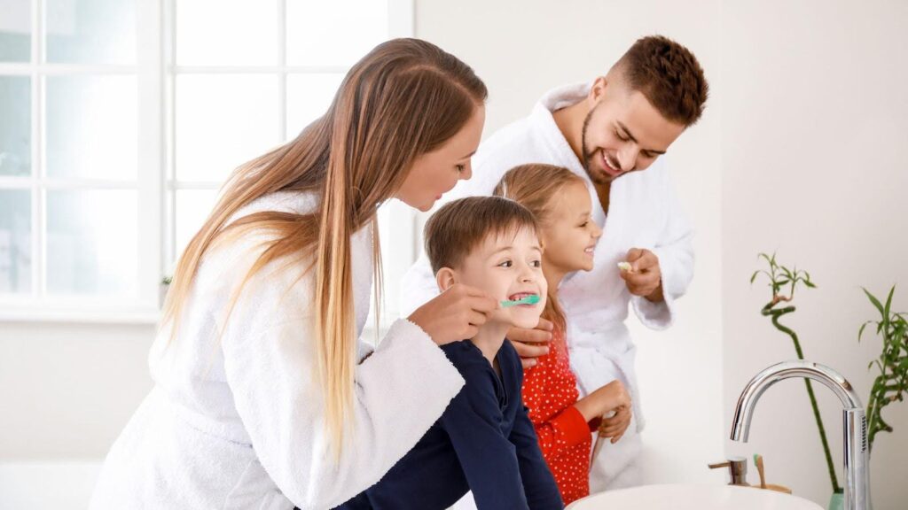 A family of four in a bathroom, with the mother helping her young son brush his teeth while the father and daughter brush their own teeth, illustrating a busy family bathroom routine.