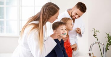 A family of four in a bathroom, with the mother helping her young son brush his teeth while the father and daughter brush their own teeth, illustrating a busy family bathroom routine.