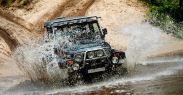 A black vehicle driving off of sand and into a body of water, creating a spray of water in the air.