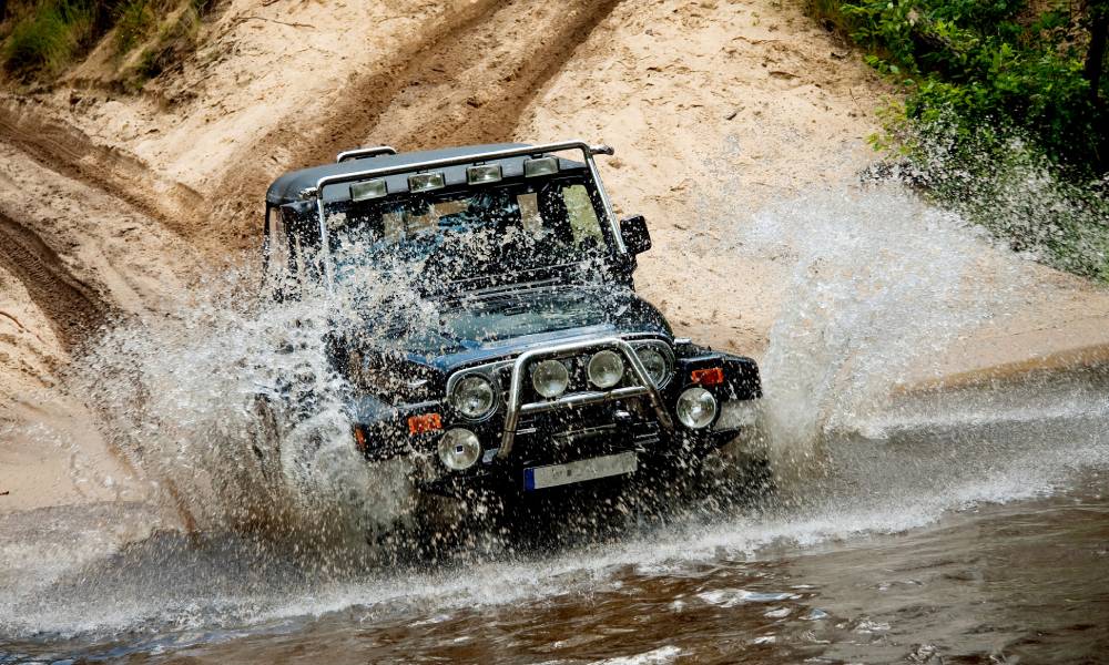 A black vehicle driving off of sand and into a body of water, creating a spray of water in the air.