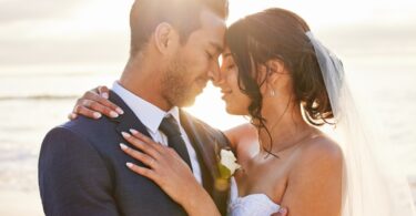 Close-up of a young couple close to each other and smiling on their wedding day with the ocean and sunset in the background.