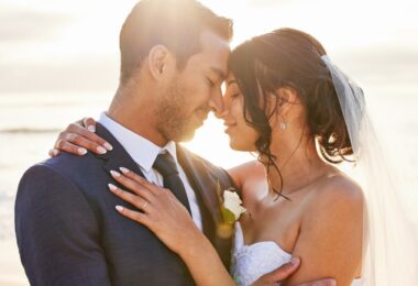 Close-up of a young couple close to each other and smiling on their wedding day with the ocean and sunset in the background.