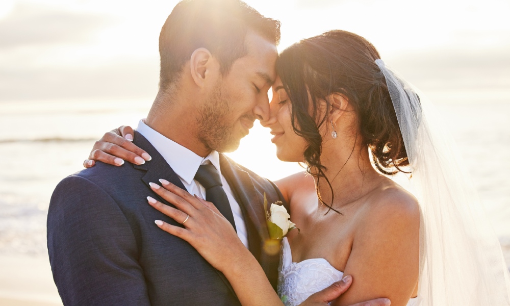 Close-up of a young couple close to each other and smiling on their wedding day with the ocean and sunset in the background.