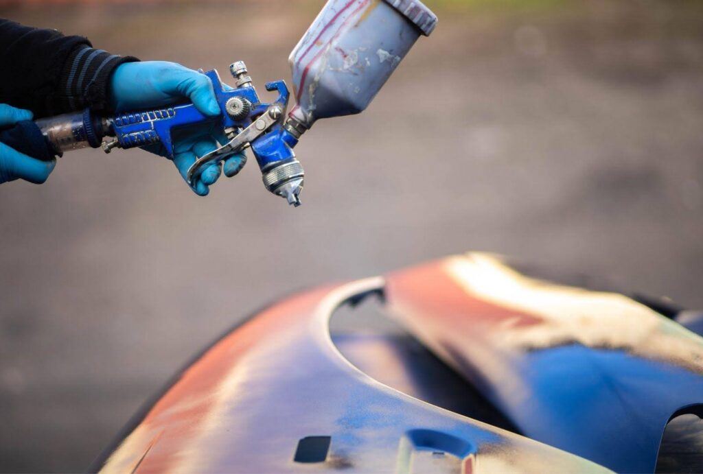 Close-up of a professional using a spray gun to apply paint correction on a car hood, illustrating the precision and expertise required for automotive paint repair.