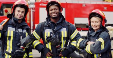 Three firefighters stand in front of a firetruck wearing their uniforms and holding oxygen masks.