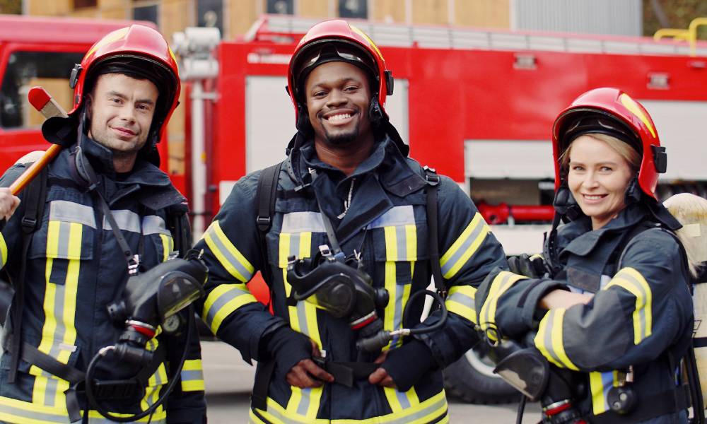 Three firefighters stand in front of a firetruck wearing their uniforms and holding oxygen masks.