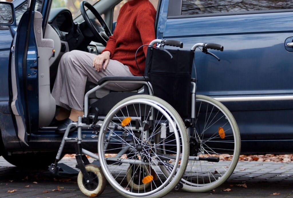 A person with mobility challenges transitions from a wheelchair into the driver's seat of a car. The open car door reveals the interior, highlighting accessible vehicle modifications designed to enhance driving safety for individuals with disabilities.