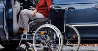 A person with mobility challenges transitions from a wheelchair into the driver's seat of a car. The open car door reveals the interior, highlighting accessible vehicle modifications designed to enhance driving safety for individuals with disabilities.