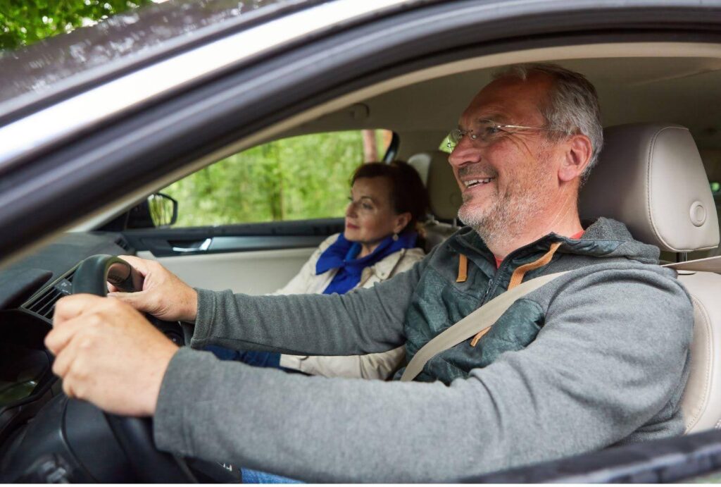 A senior man smiling while driving a car, with a senior woman passenger looking relaxed beside him. The interior of the car is well-lit, showing the man holding the steering wheel and both wearing casual attire, capturing a moment of joyful travel in a comfortable vehicle setting.