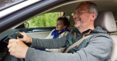 A senior man smiling while driving a car, with a senior woman passenger looking relaxed beside him. The interior of the car is well-lit, showing the man holding the steering wheel and both wearing casual attire, capturing a moment of joyful travel in a comfortable vehicle setting.