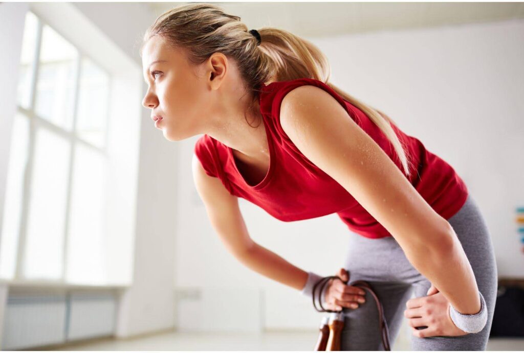 A young female athlete leaning over and resting after a strenuous workout, wearing a red top and grey leggings, demonstrating fatigue and recovery in a bright, airy indoor setting.