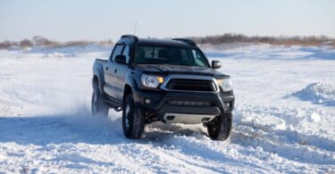 A black truck drives through the snow in winter. The sky is gray and and the truck kicks up snow as it drives.