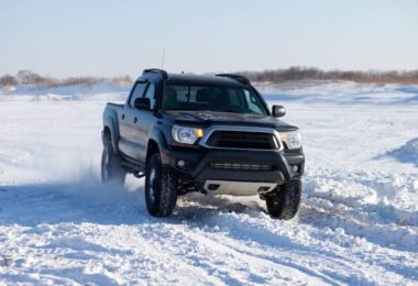 A black truck drives through the snow in winter. The sky is gray and and the truck kicks up snow as it drives.