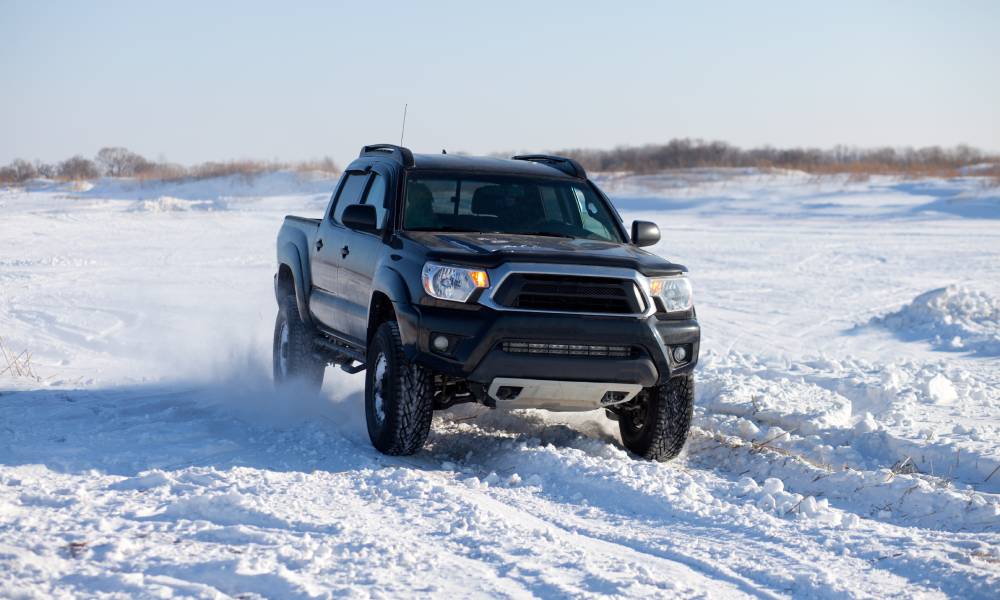 A black truck drives through the snow in winter. The sky is gray and and the truck kicks up snow as it drives.