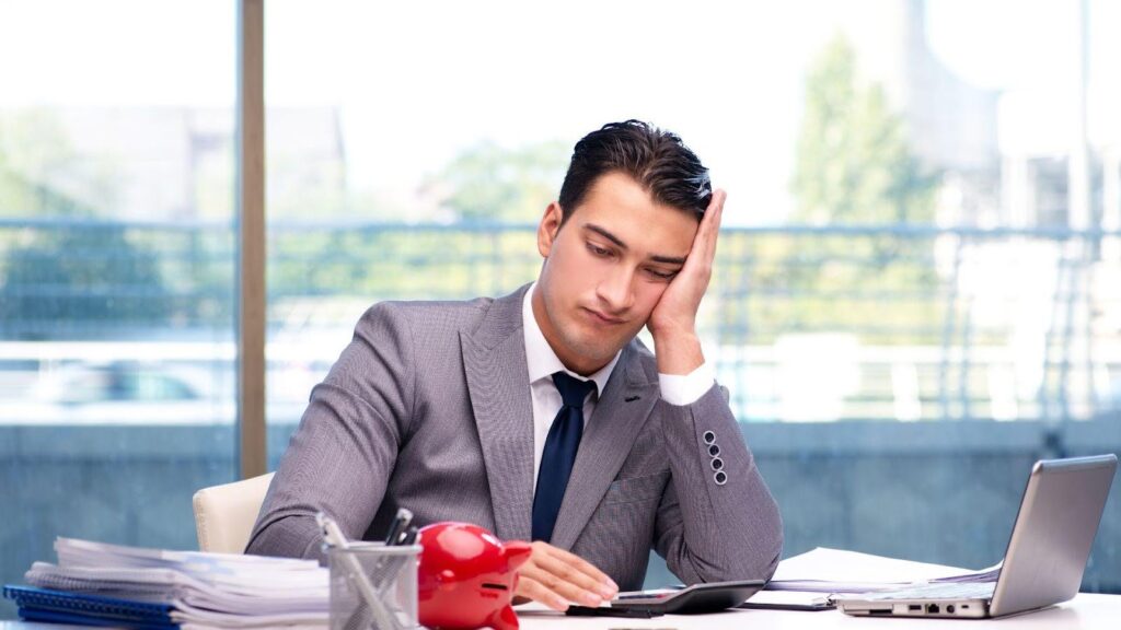 Businessman sitting at a desk looking stressed with laptops and paperwork around