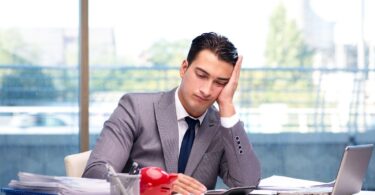 Businessman sitting at a desk looking stressed with laptops and paperwork around