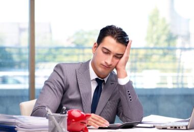 Businessman sitting at a desk looking stressed with laptops and paperwork around