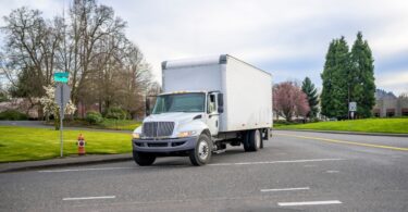 A small compact semi truck with a cube box trailer is driving on an urban city street, surrounded by blooming trees.