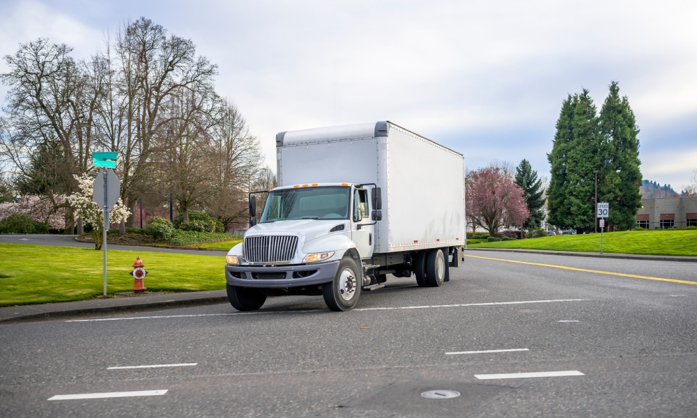 A small compact semi truck with a cube box trailer is driving on an urban city street, surrounded by blooming trees.