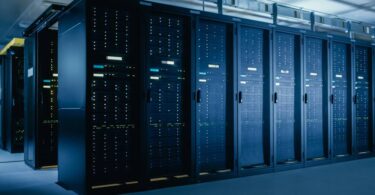 Long rows of black server racks fill the room of a large data center. Low lighting can be seen overhead.