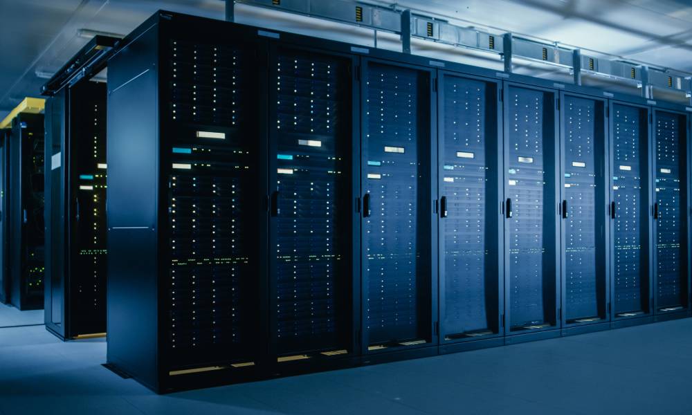 Long rows of black server racks fill the room of a large data center. Low lighting can be seen overhead.