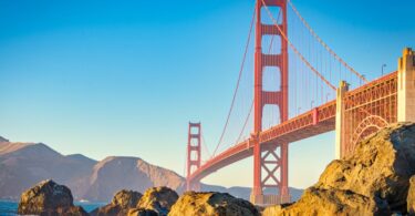 The Golden Gate Bridge framed by rocks at the water level. The scenery and bridge are painted in warm hues as the sun sets.