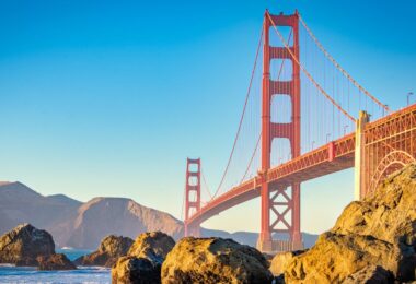 The Golden Gate Bridge framed by rocks at the water level. The scenery and bridge are painted in warm hues as the sun sets.