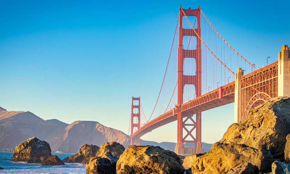 The Golden Gate Bridge framed by rocks at the water level. The scenery and bridge are painted in warm hues as the sun sets.