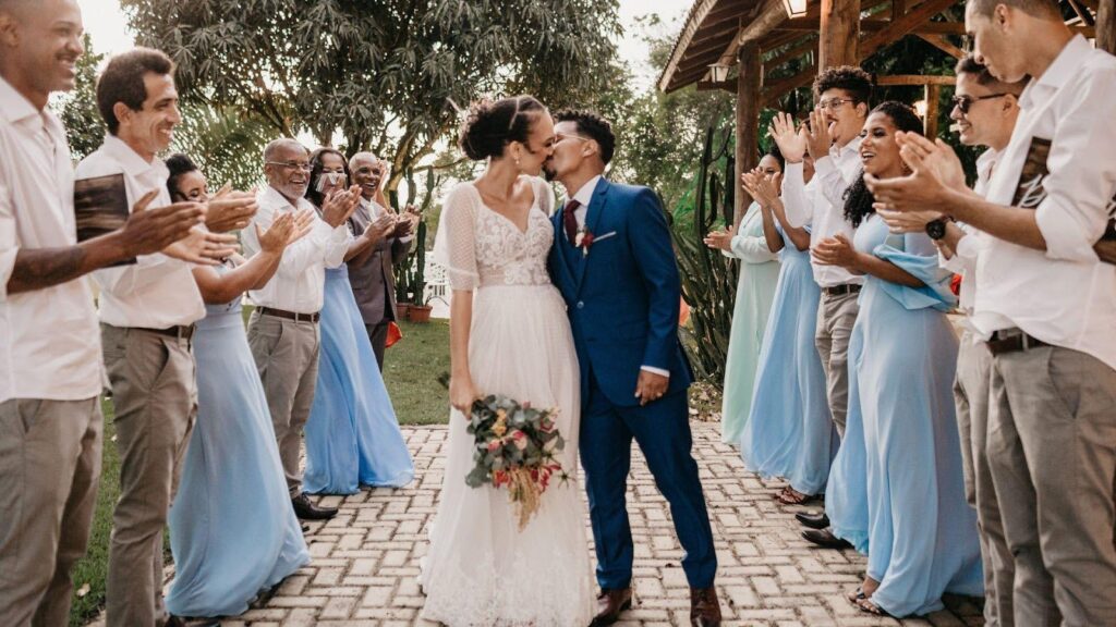 A bride and groom kissing, surrounded by cheering guests at an outdoor wedding celebration, capturing a moment of joy and community support. 