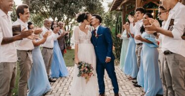 A bride and groom kissing, surrounded by cheering guests at an outdoor wedding celebration, capturing a moment of joy and community support.